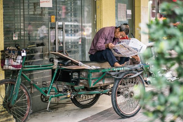 An older Chinese man takes a break on his three-wheeled bicycle and intently reads the daily newspaper.