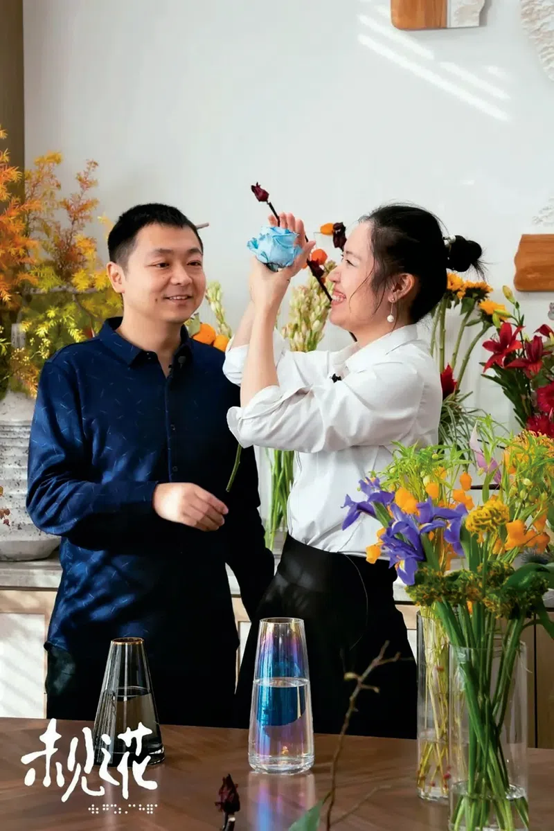 Xia Jia examines a blue flower's petals as she lets out a big smile as a man in a blue shirt watches her enjoy the flowers.