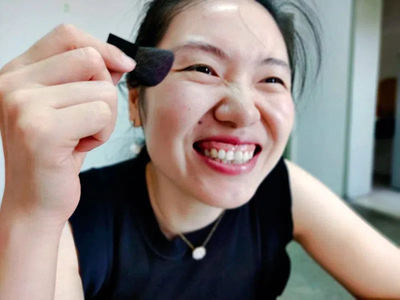 Xiao Jie, wearing a navy outfit happily smiles in front of the camera while holding a small, black makeup brush in front of her face.