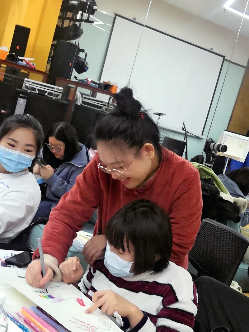 Pei Yujia smiles while assisting a young student with her assignment while she wears a brownish-red sweater.