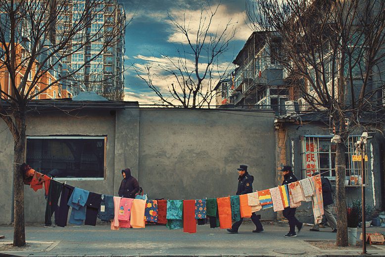 A vendor hangs garment for sales on a street that used to have several clothing stores, now all walled up; she pretends to be a regular passerby as security guards walk past